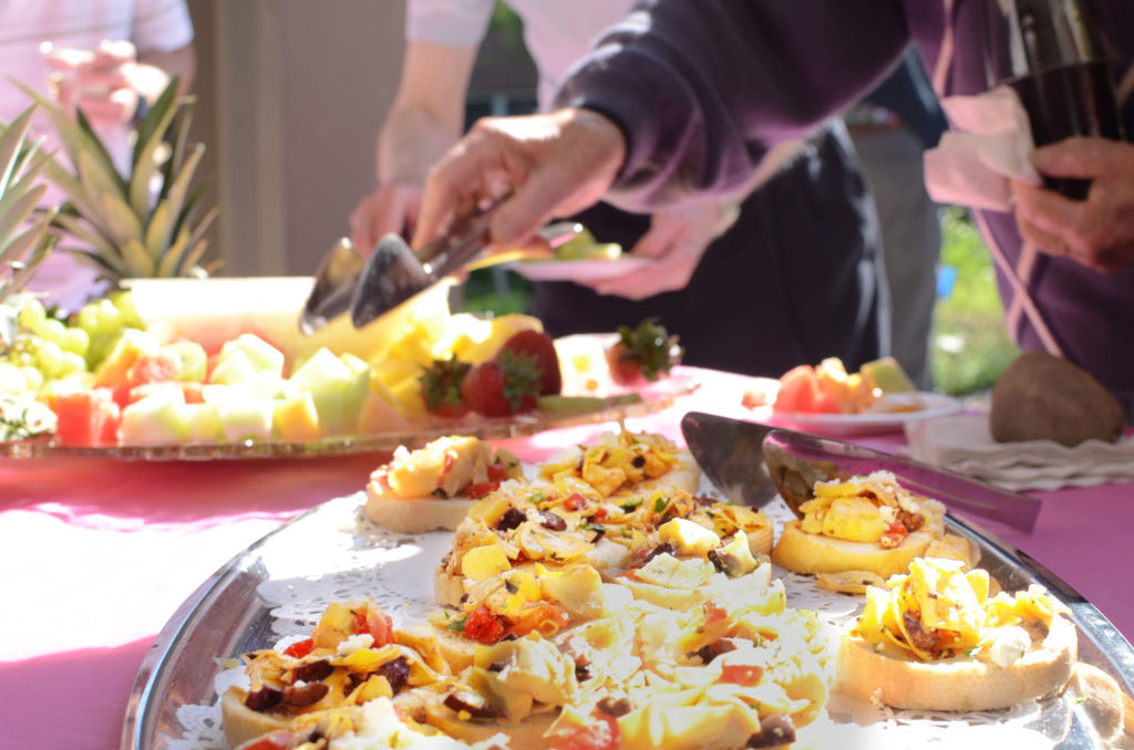 A senior resident uses tongs to pick up fresh cut fruit, hors d’oeuvres and appetizers from an outside table